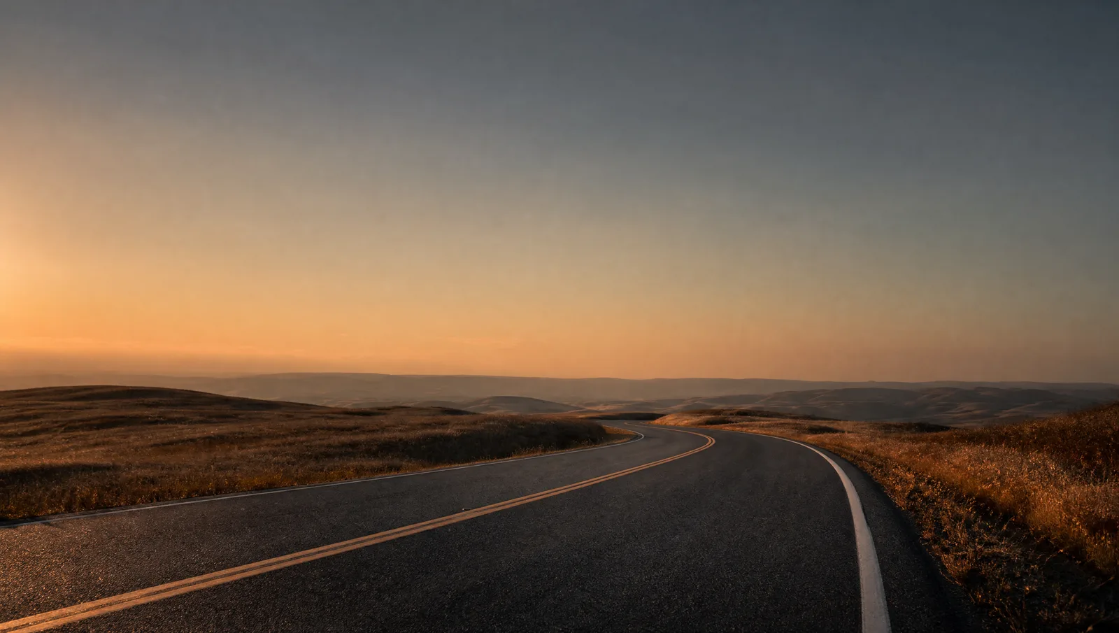 An empty rural road curves through open hills under a dusky amber-to-blue sky, the asphalt catching low raking light, with no cars, signs, or buildings in sight.